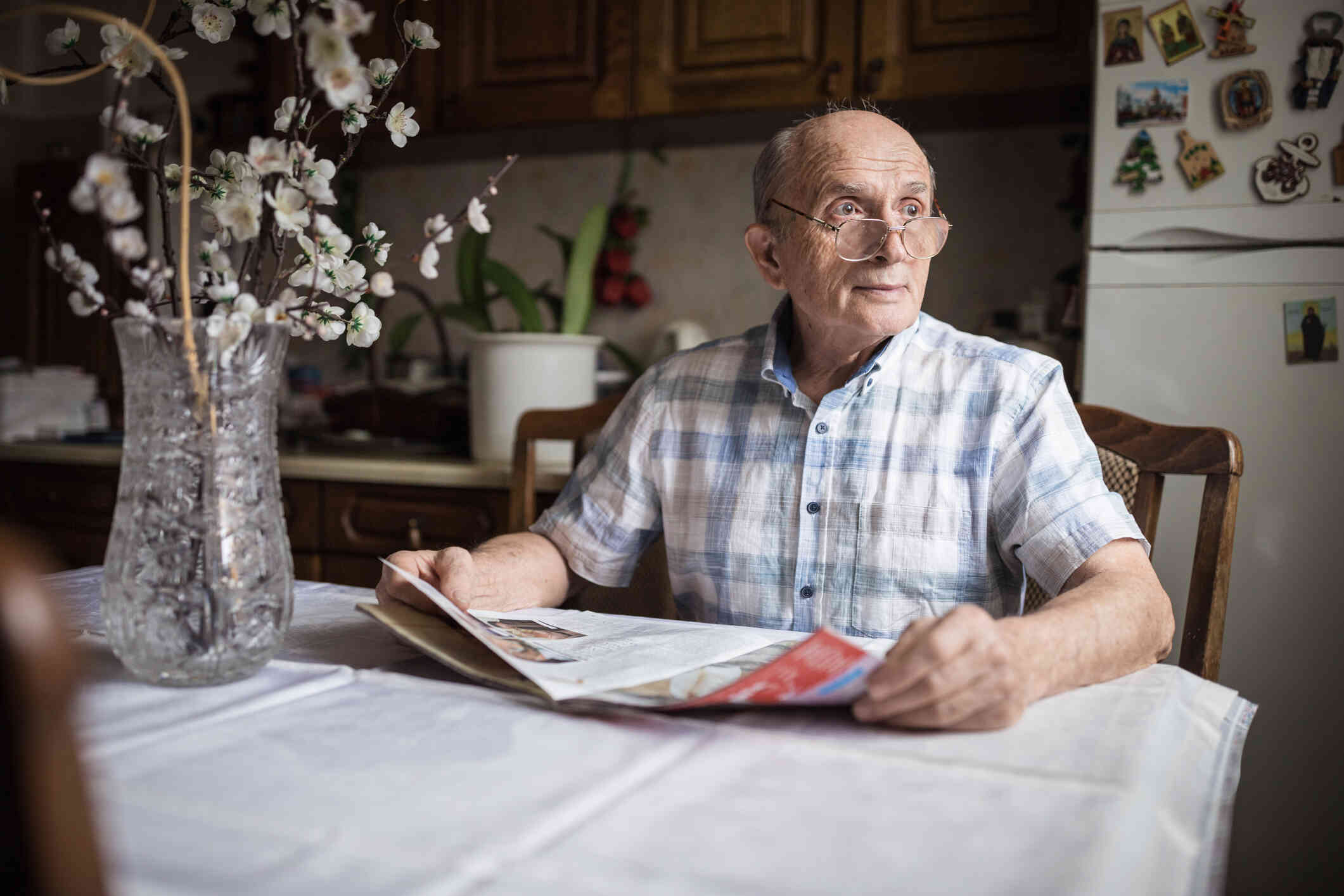 An elderly man sits at the kitchen table with a magazine and gazes off.
